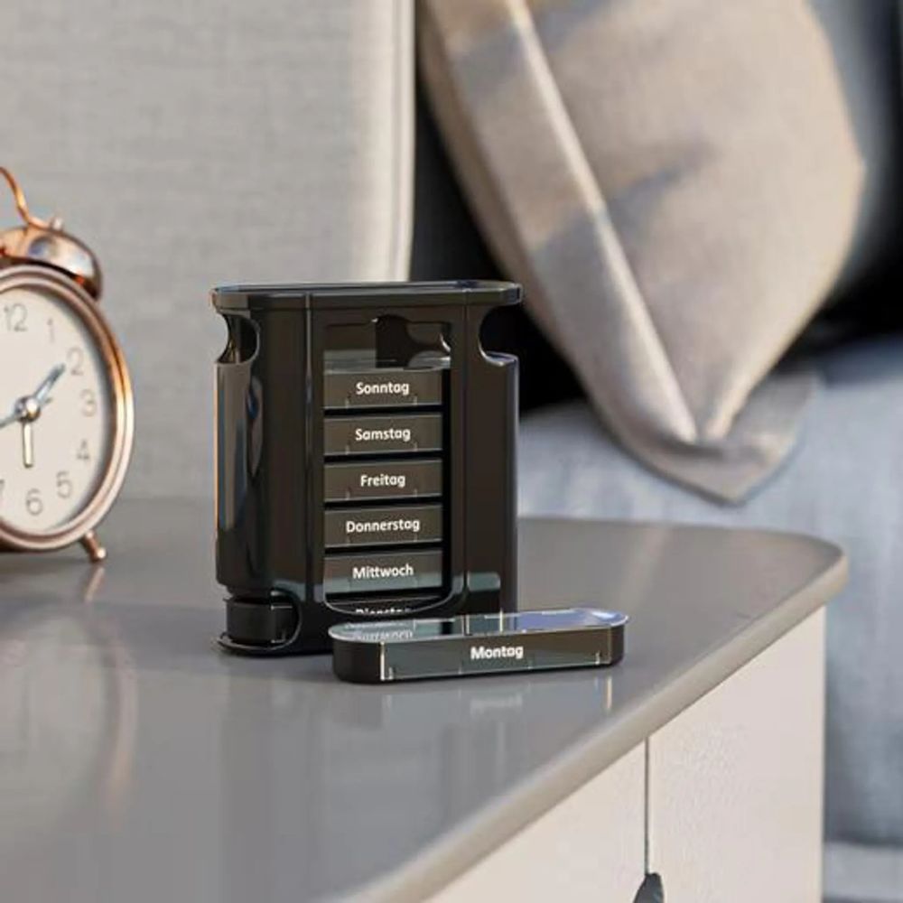 Schwarze Tablettenbox auf einer grauen Oberfläche. Ein herausgenommenes Fach mit der Aufschrift Montag. Im Hintergrund eine Uhr und ein Kissen.