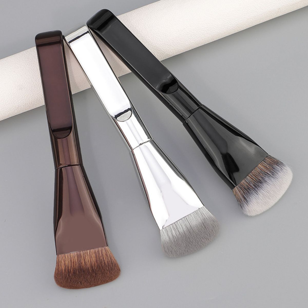 Drei flache Make-up-Pinsel mit glänzenden Griffen in Braun, Silber und Schwarz.