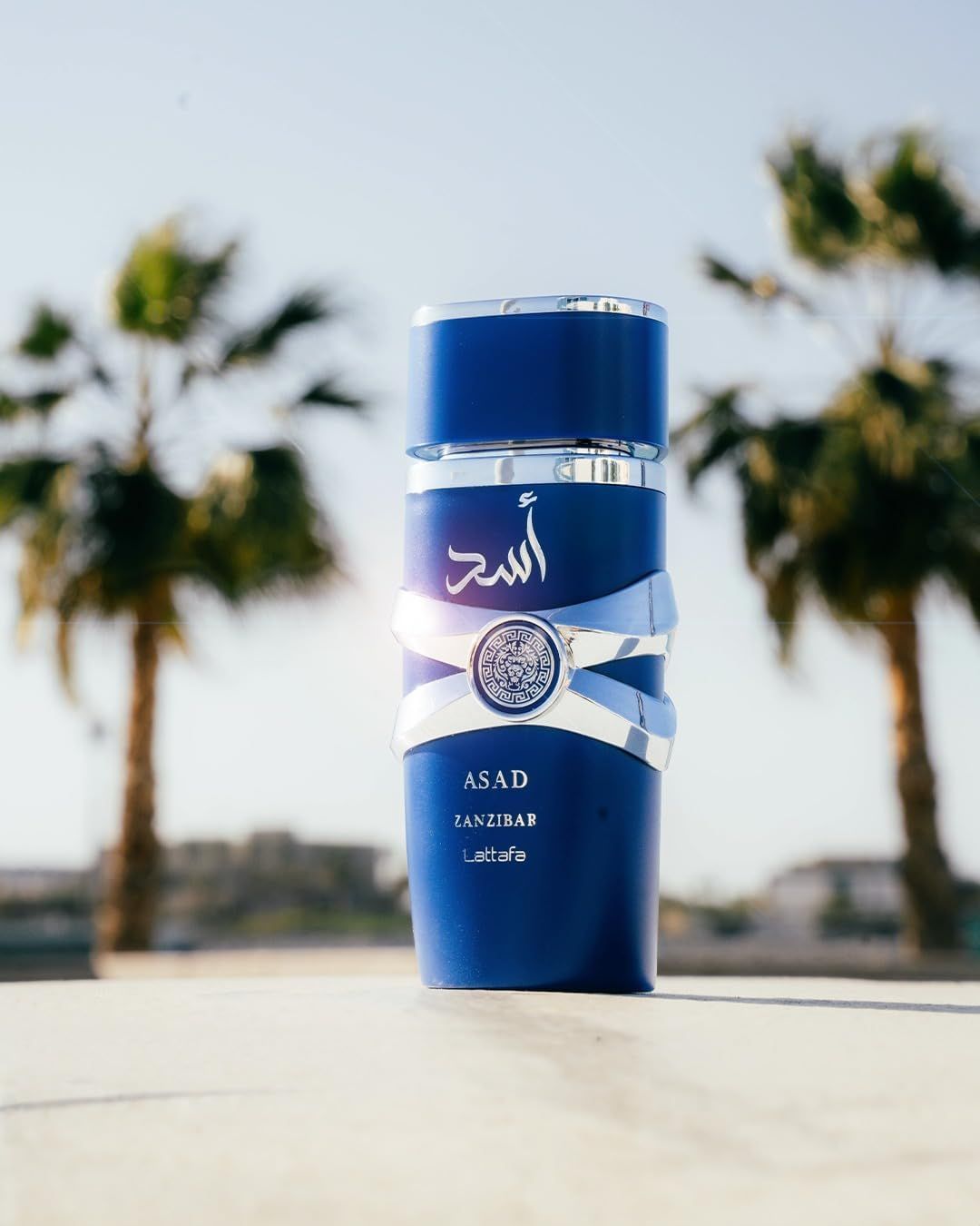 Blauer Flakon mit silbernen Bändern und Logo. Aufschrift: ASAD, ZANZIBAR, Lattafa. Palmen im Hintergrund, sonniges Wetter.