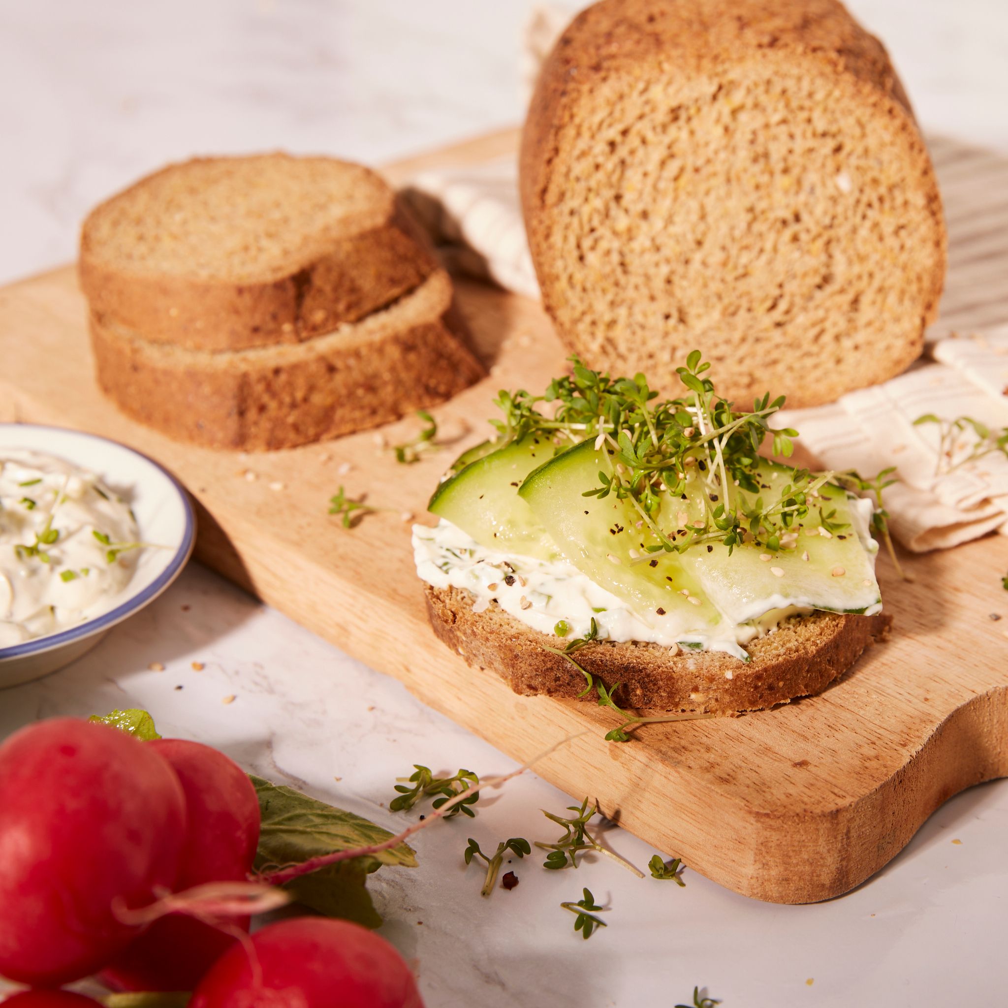 Geschnittenes Brot auf Holzbrett. Belegtes Brot mit Gurke und Kresse. Radieschen und Dip daneben.