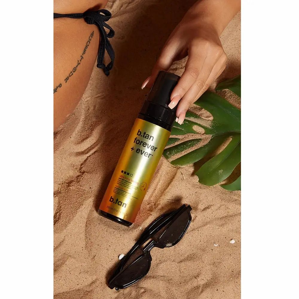 Selbstbräunungsmousse-Flasche auf Sand. Schwarz-goldene Verpackung. Text: b.tan forever + ever. Sonnenbrille und Blatt daneben. Hand hält Flasche.