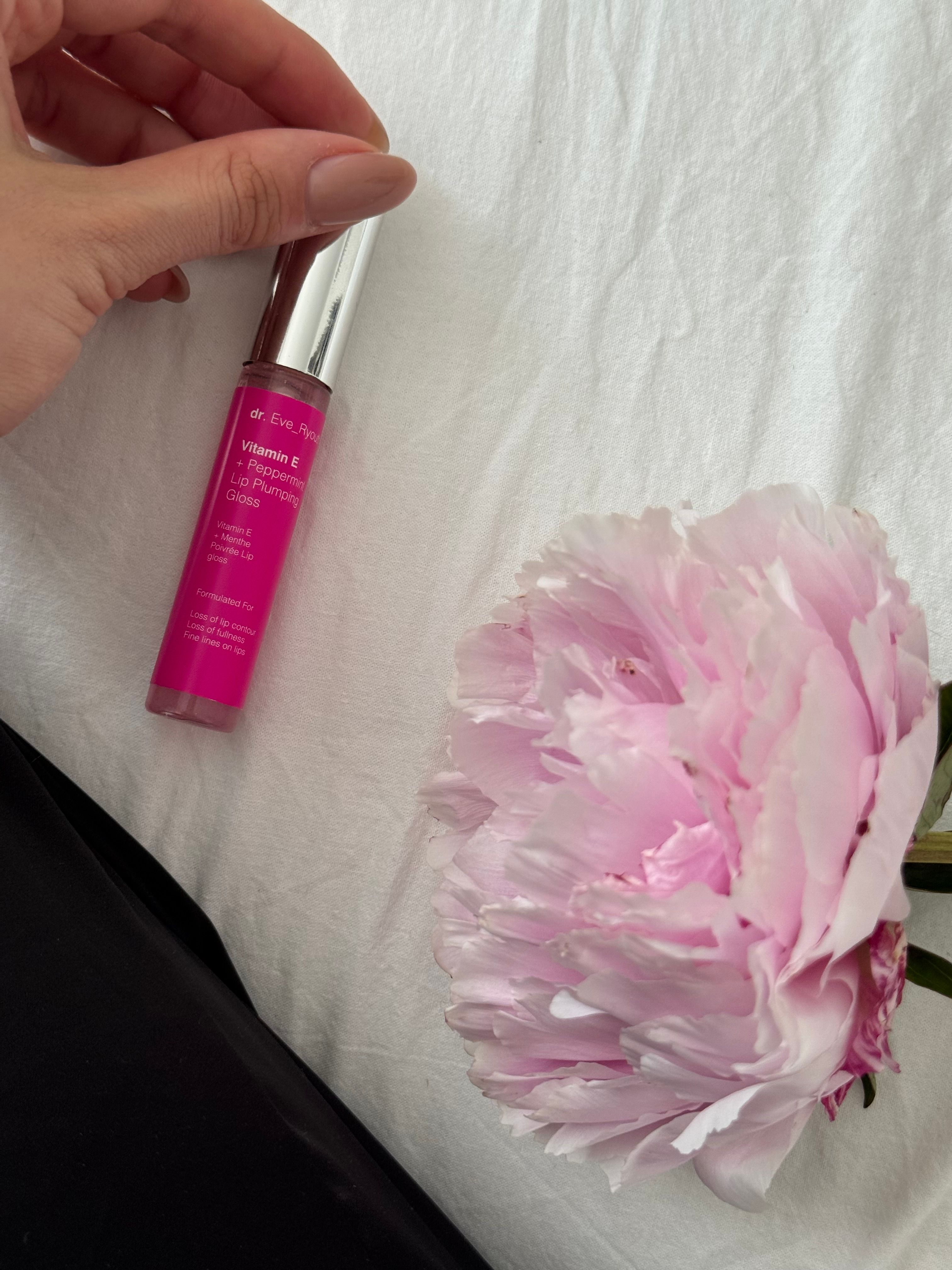 Eine Lippenfüller-Tube neben einer Blüte. Rosa Flüssigkeit in durchsichtigem Behälter mit silbernem Deckel. Aufschrift: Dr. Eve_Ryouth, Vitamin E + Pfefferminz.