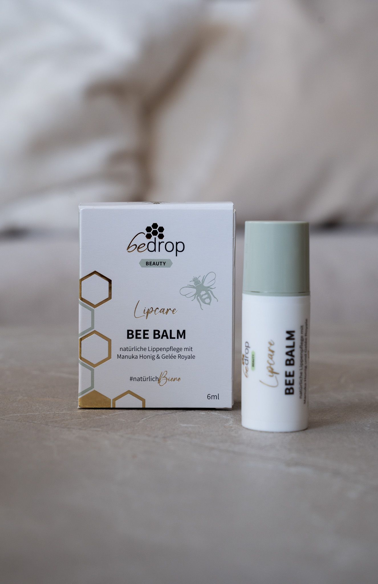 Lippenbalsam und Verpackung auf hellem Stoff. Aufschrift: BEE BALM, Lipcare, bedrop. Verpackung mit Bienen-Logo.