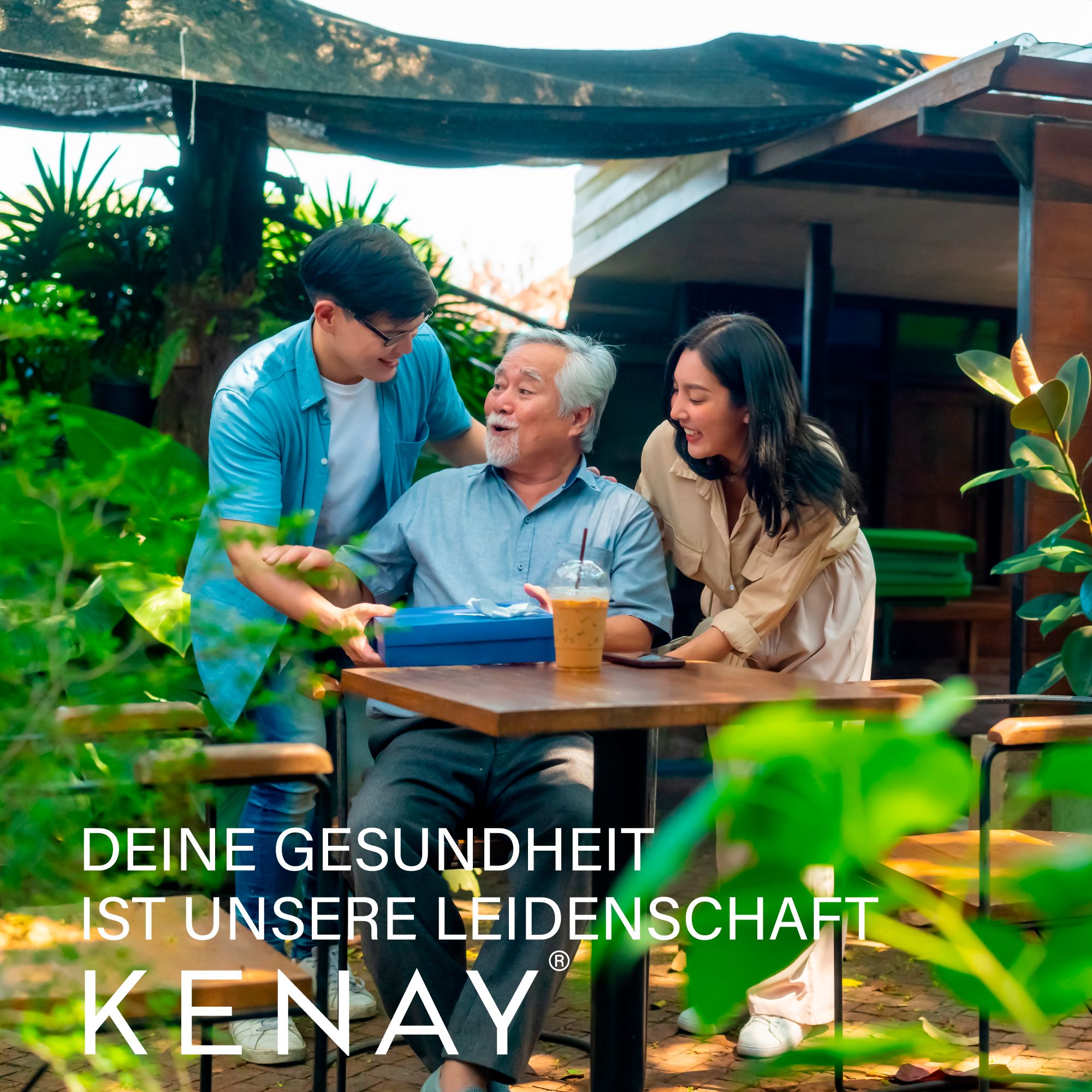 Älterer Mann erhält Geschenk von jungem Paar. Text: DEINE GESUNDHEIT IST UNSERE LEIDENSCHAFT. KENAY®-Logo.