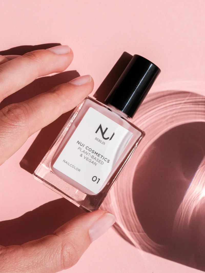 Hand hält NUI Cosmetics Nagellack Rosé. Rechteckige Glasflasche mit schwarzem Deckel. Etikett mit Logo, Produktname und Farbnummer 01. Rosa Hintergrund.