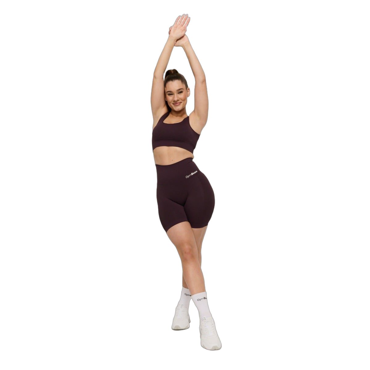 Frau in dunkelrotem Sport-BH und Radlerhose, hebt die Arme. Nahtloses Design, breite Träger.
