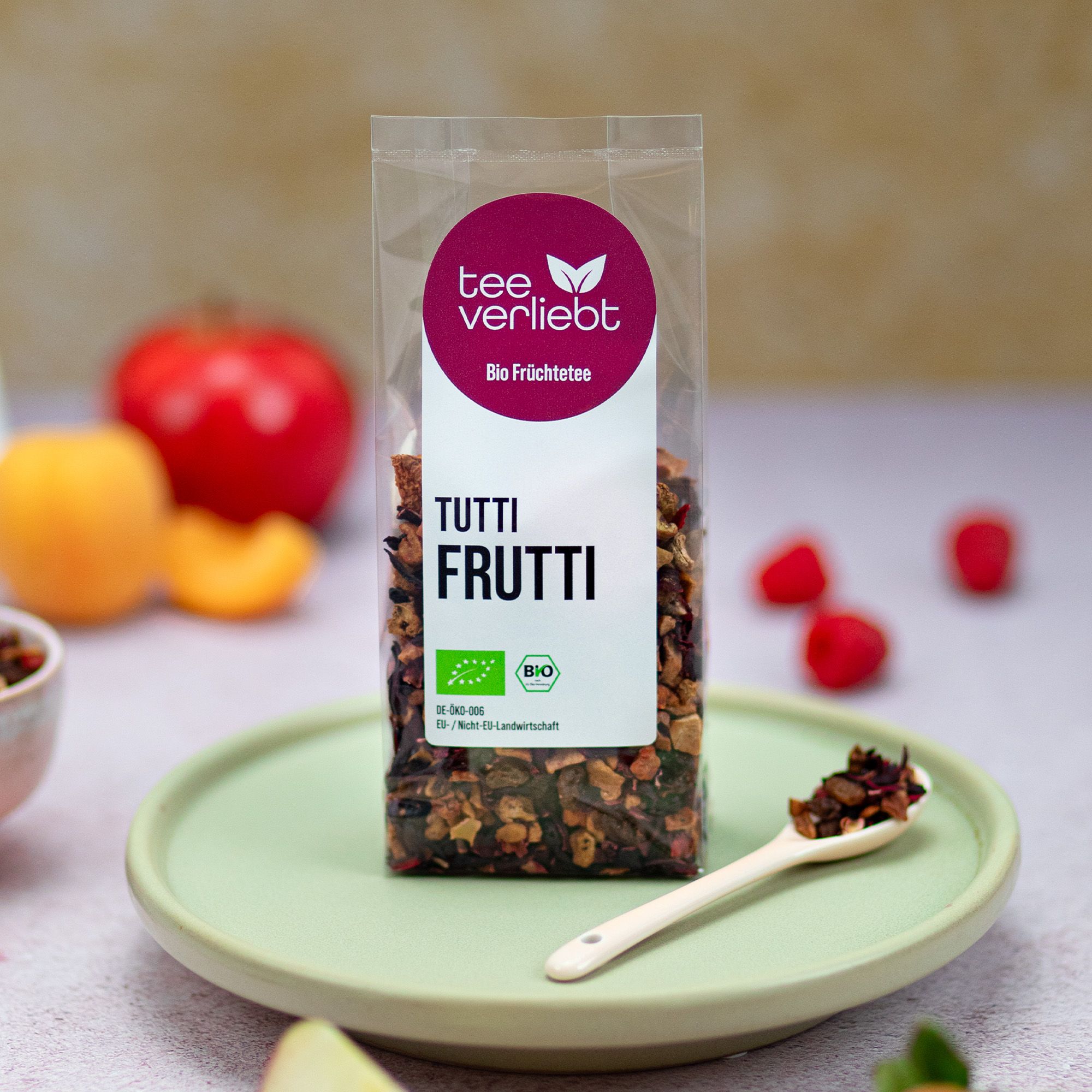 Verpackung mit losem Früchtetee auf einem Teller. Logo 'tee verliebt' und Text 'TUTTI FRUTTI'. Löffel mit Tee daneben. Früchte im Hintergrund.
