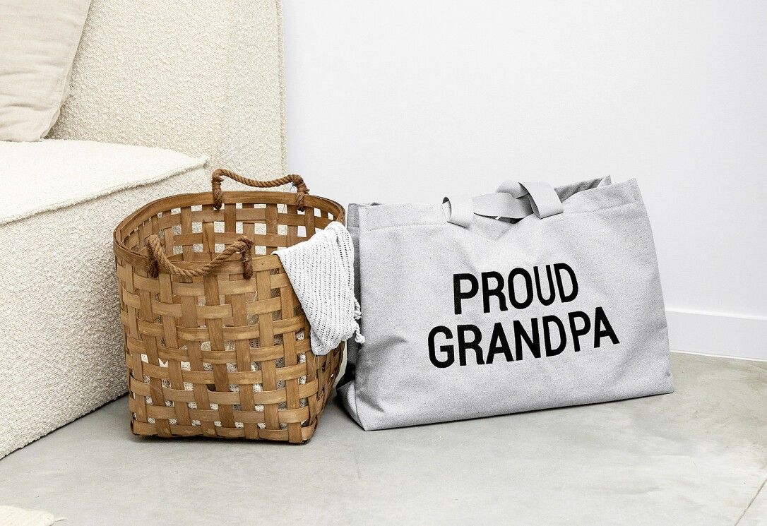 Graue Leinwandtasche mit Aufschrift "PROUD GRANDPA" neben einem Weidenkorb. Beide stehen auf dem Boden.