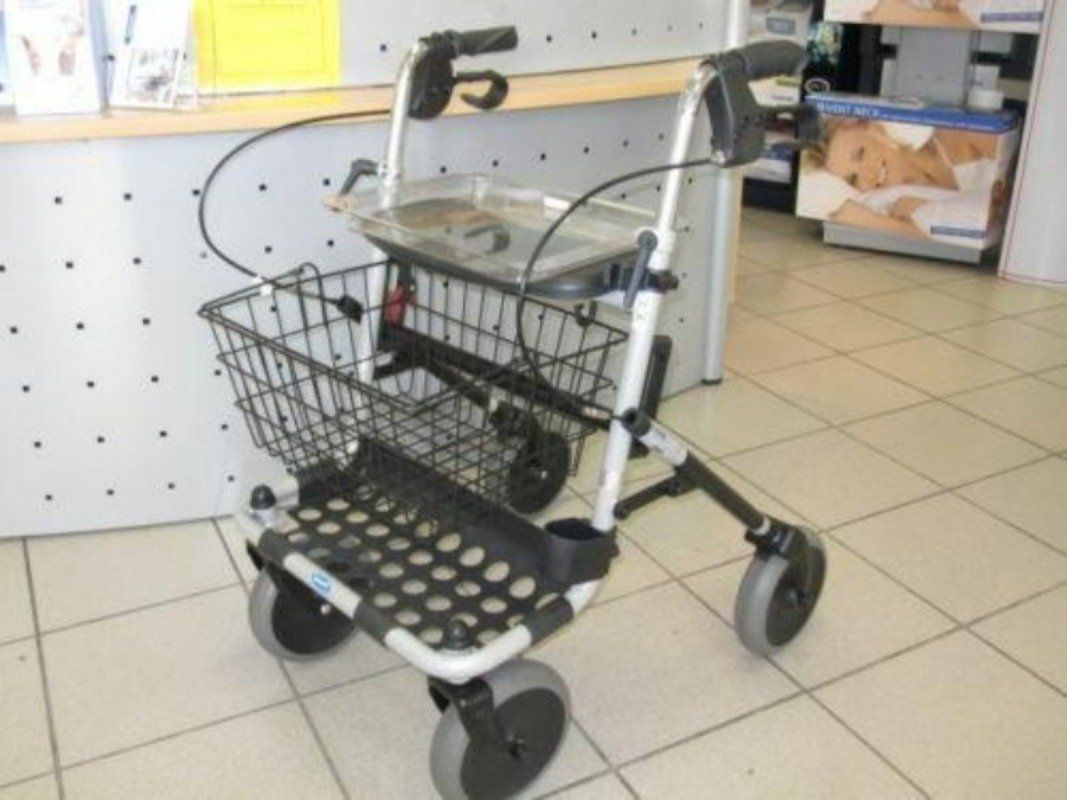 Rollator mit Korb, Tablett und vier Rädern. Silberfarbener Rahmen, schwarzer Korb und Sitzfläche. Graue Räder. Im Hintergrund ein Geschäft.