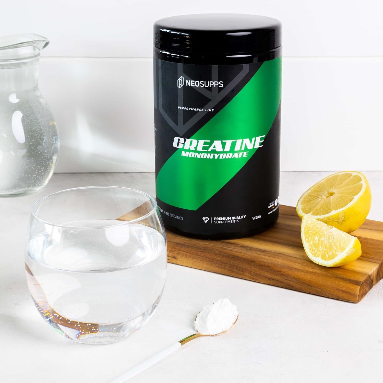 Schwarze Dose mit grünem Etikett. Aufschrift: Creatine Monohydrate. Marke: Neosupps. Neben Glas Wasser, Zitrone und Löffel mit Pulver.