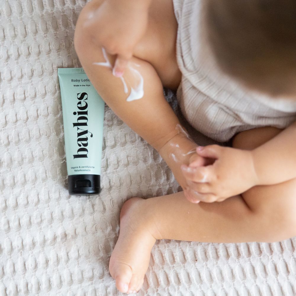 Eine hellgrüne Tube 'baybies Baby Lotion' neben einem Babybein, auf das Lotion aufgetragen wird.