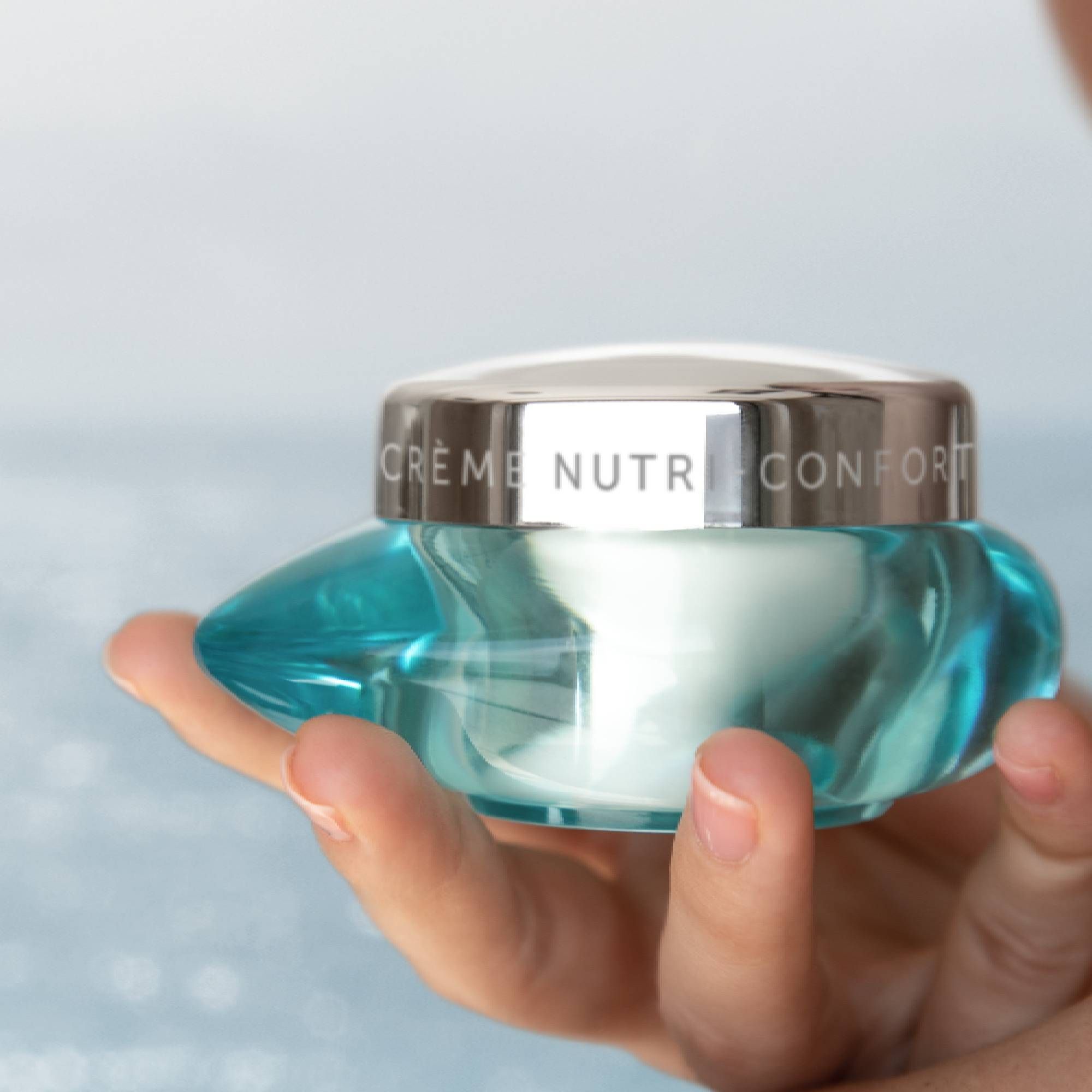 Türkisfarbener Tiegel mit silbernem Deckel, gehalten von einer Hand. Aufschrift: Crème Nutri-Confort. Produktname nicht enthalten.