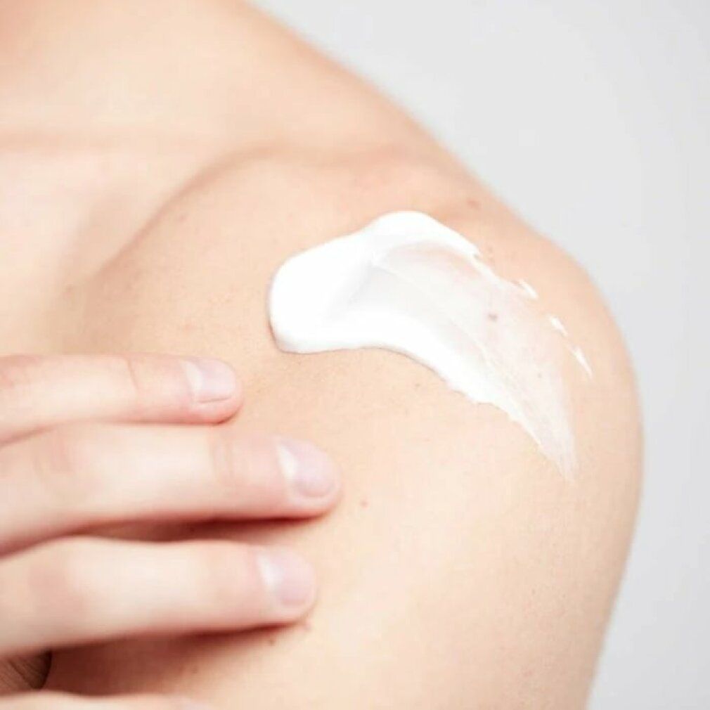 Person trägt cremefarbene Creme auf die Schulter auf. Hand berührt die Creme.