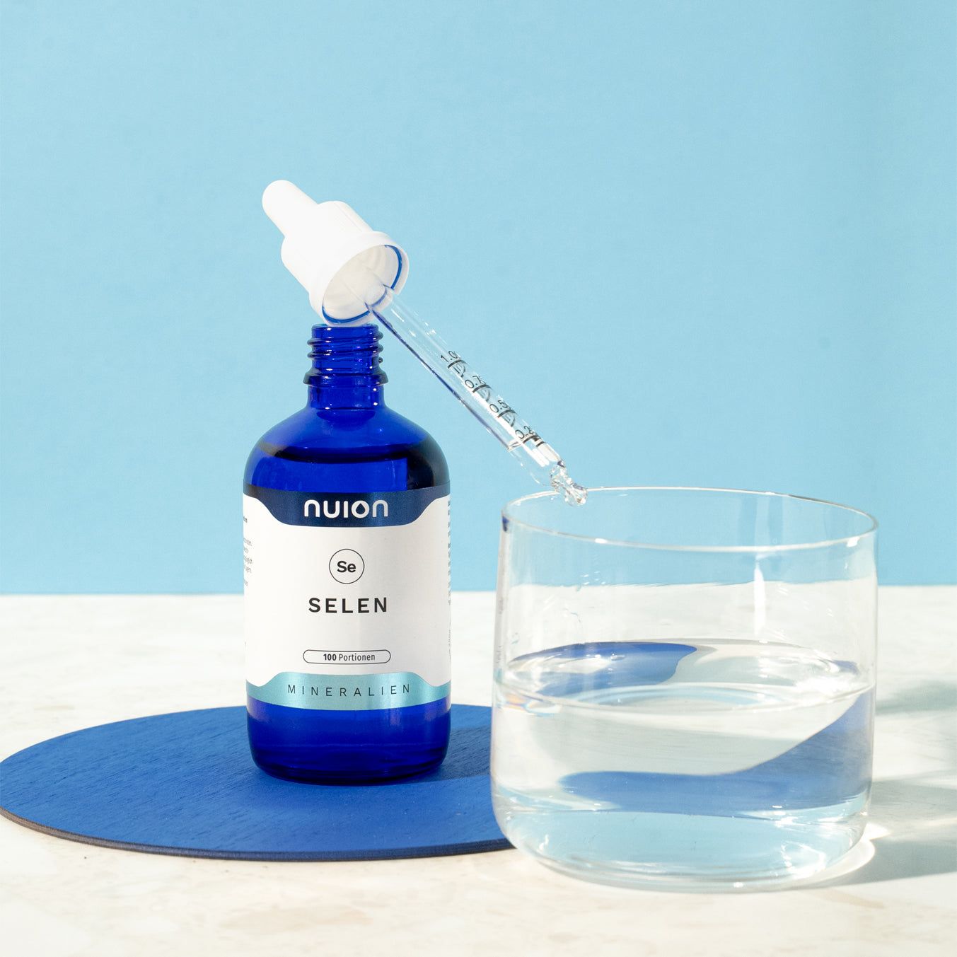 Nuion Selen Tropfenflasche mit Pipette, 100 Portionen, neben einem Glas Wasser.