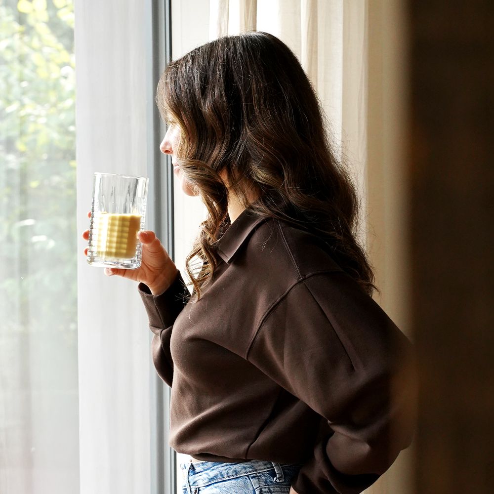 Frau hält ein Glas mit gelbem Getränk. Sie steht vor einem Fenster.