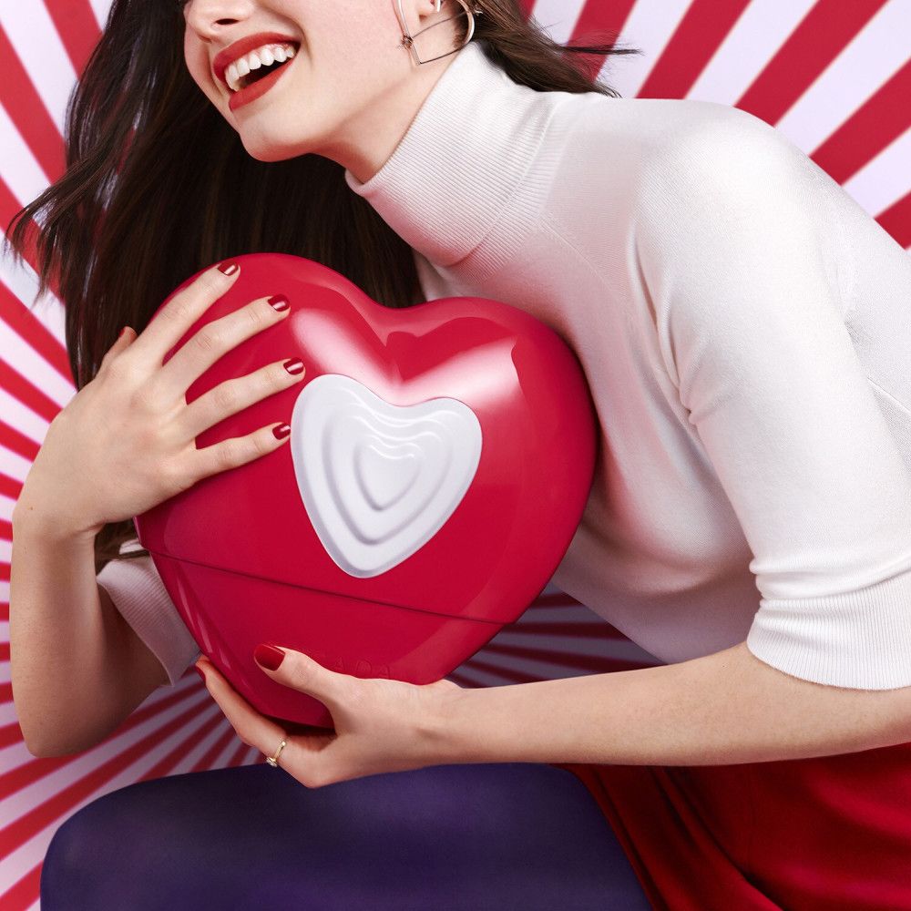 Eine Frau hält einen roten Herzflakon mit weißem Herz. Sie trägt ein weißes Oberteil und hat rote Lippen und Fingernägel.