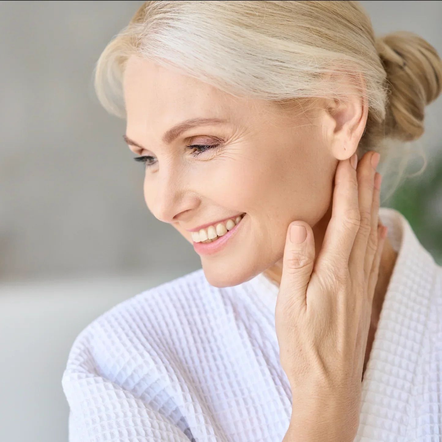 Frau mit Bademantel lächelt. Hält Hand an Hals. Helle Umgebung.