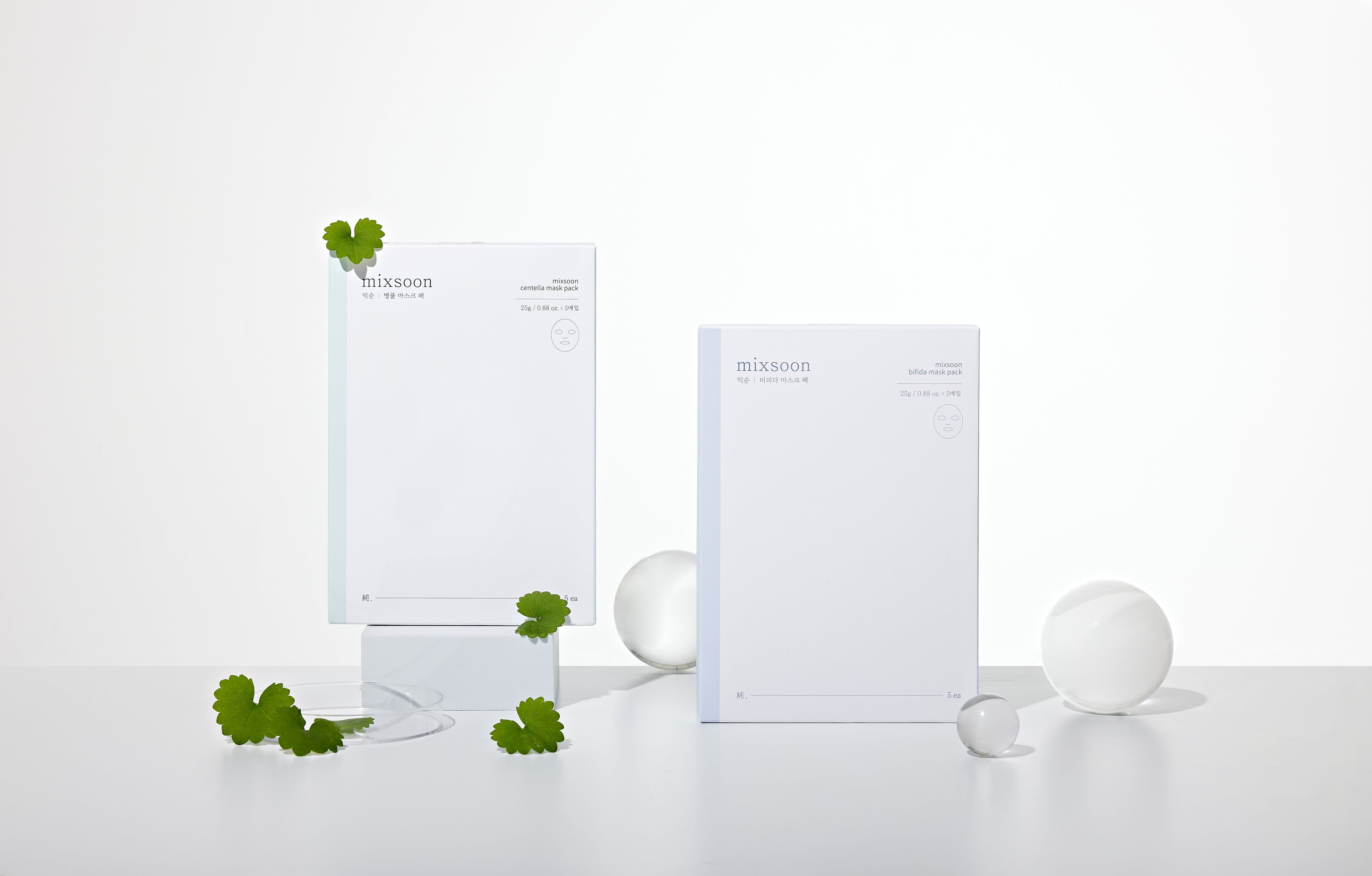 Zwei Mixsoon Centella Mask Pack-Verpackungen. Weiße, rechteckige Pakete. Daneben grüne Blätter und transparente Kugeln.