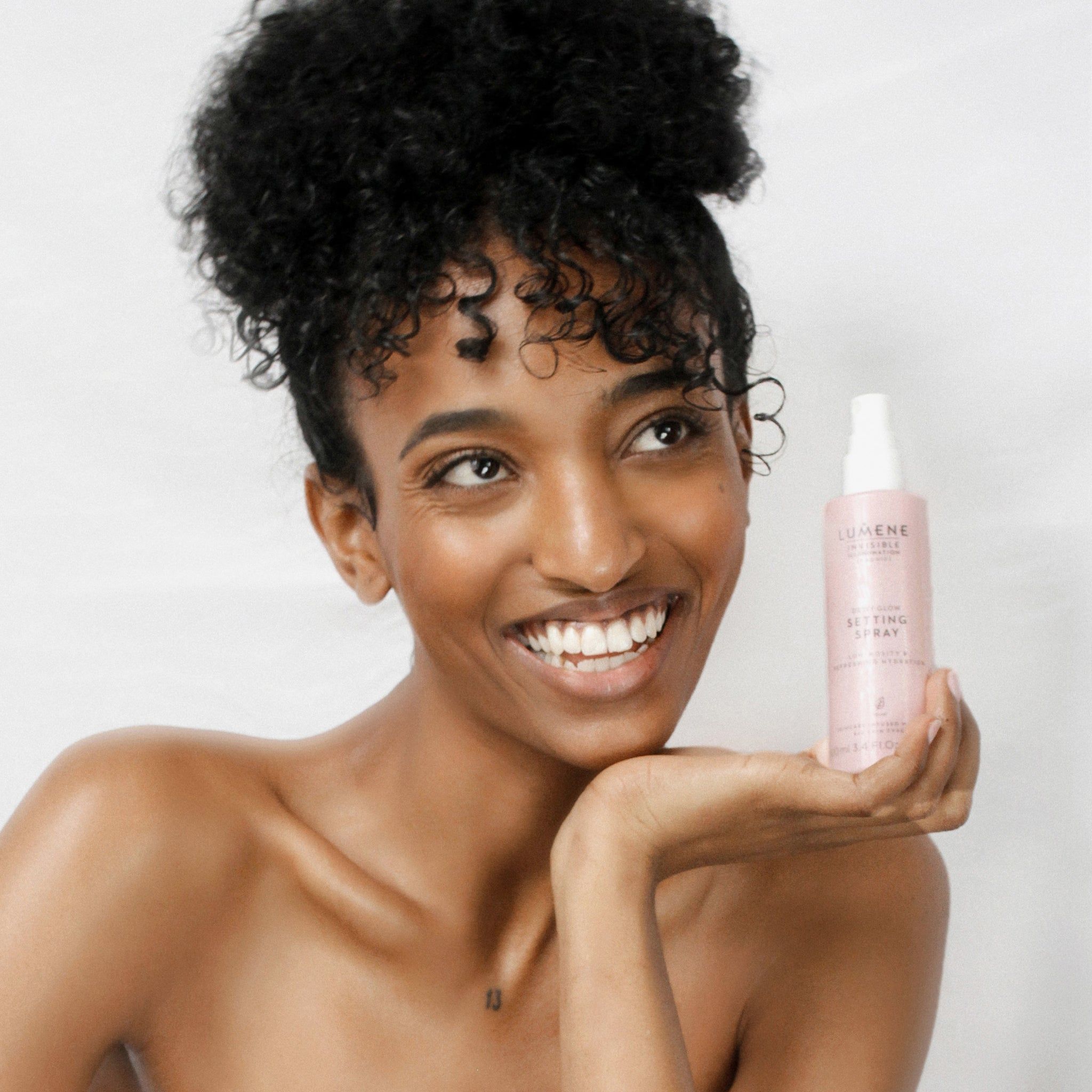 Frau hält rosa Sprühflasche. Aufschrift: Lumene INVISIBLE ILLUMINATION Dewy Glow Setting Spray. Frau lächelt. Hintergrund: weiß.