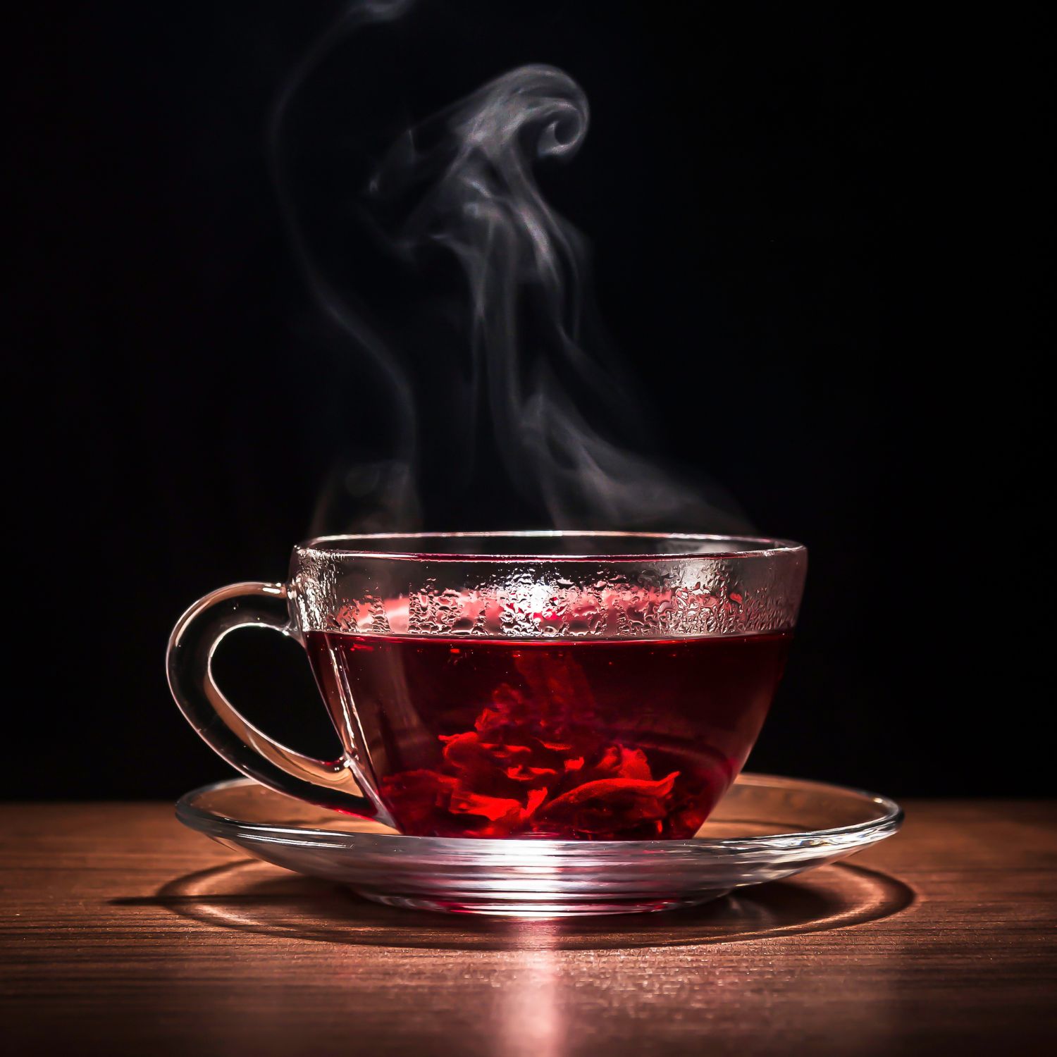 Glas-Teetasse mit rotem Getränk. Dampf steigt auf. Auf Untertasse. Auf Holzoberfläche, dunkler Hintergrund.