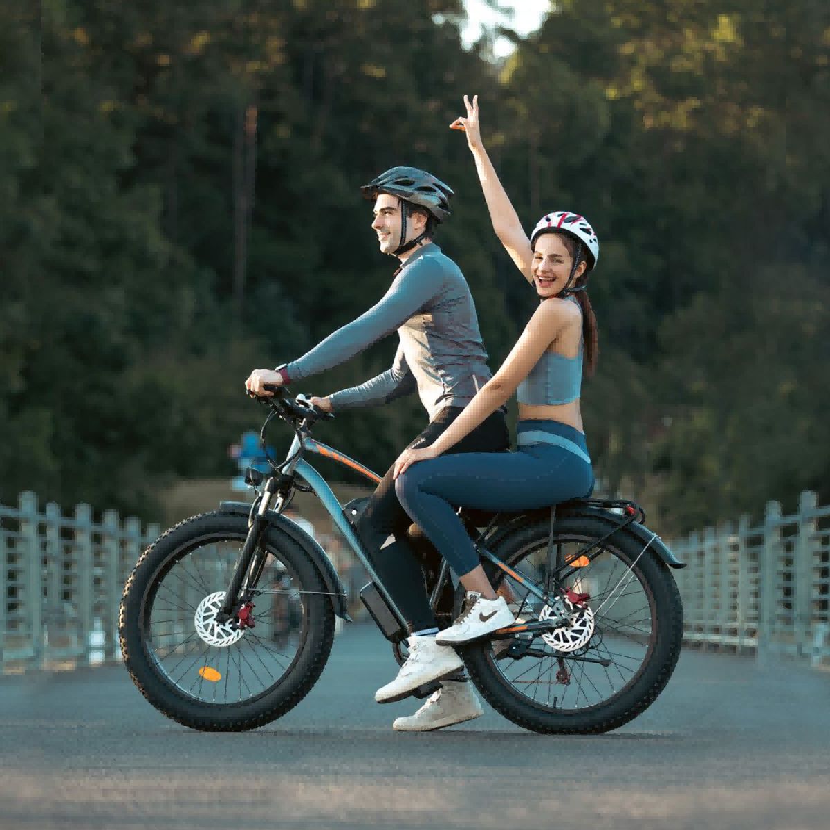 Zwei Personen fahren auf einem grauen Elektro-Fatbike. Sie tragen Helme. Hintergrund: Bäume und Brücke.