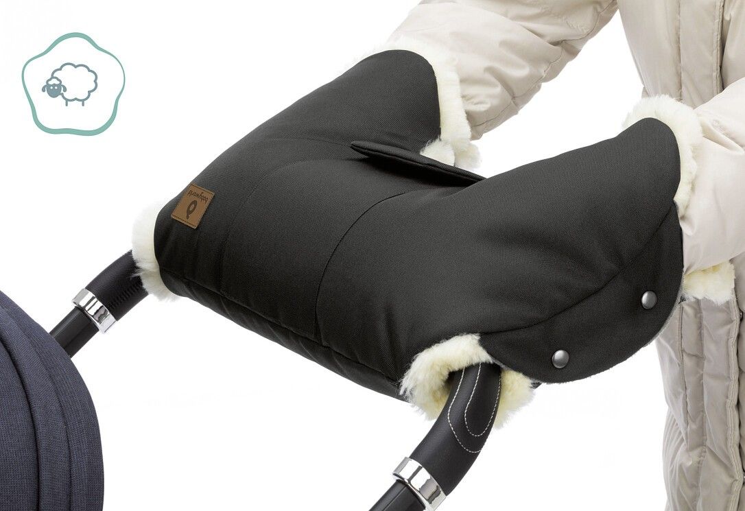 Schwarzer Handwärmer mit Lammfellbesatz an einem Kinderwagen befestigt. Logo-Etikett sichtbar. Person hält den Handwärmer.
