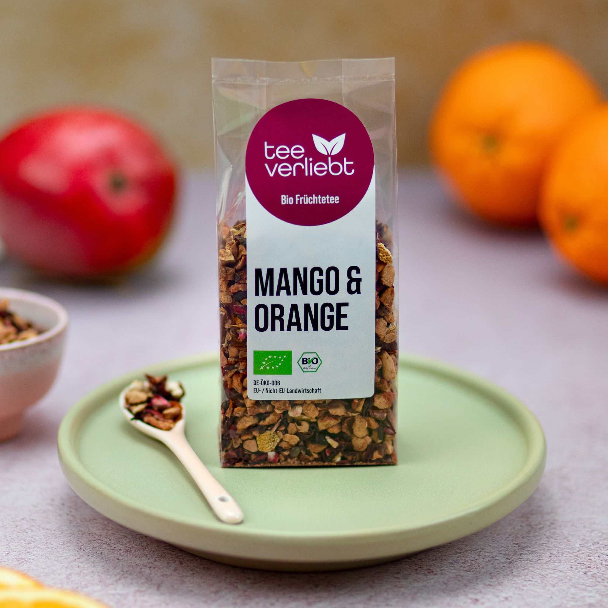 Teebeutel auf Teller mit Löffel. Mango, Orangen im Hintergrund. Aufdruck: "tee verliebt", "Mango & Orange", Bio-Siegel.