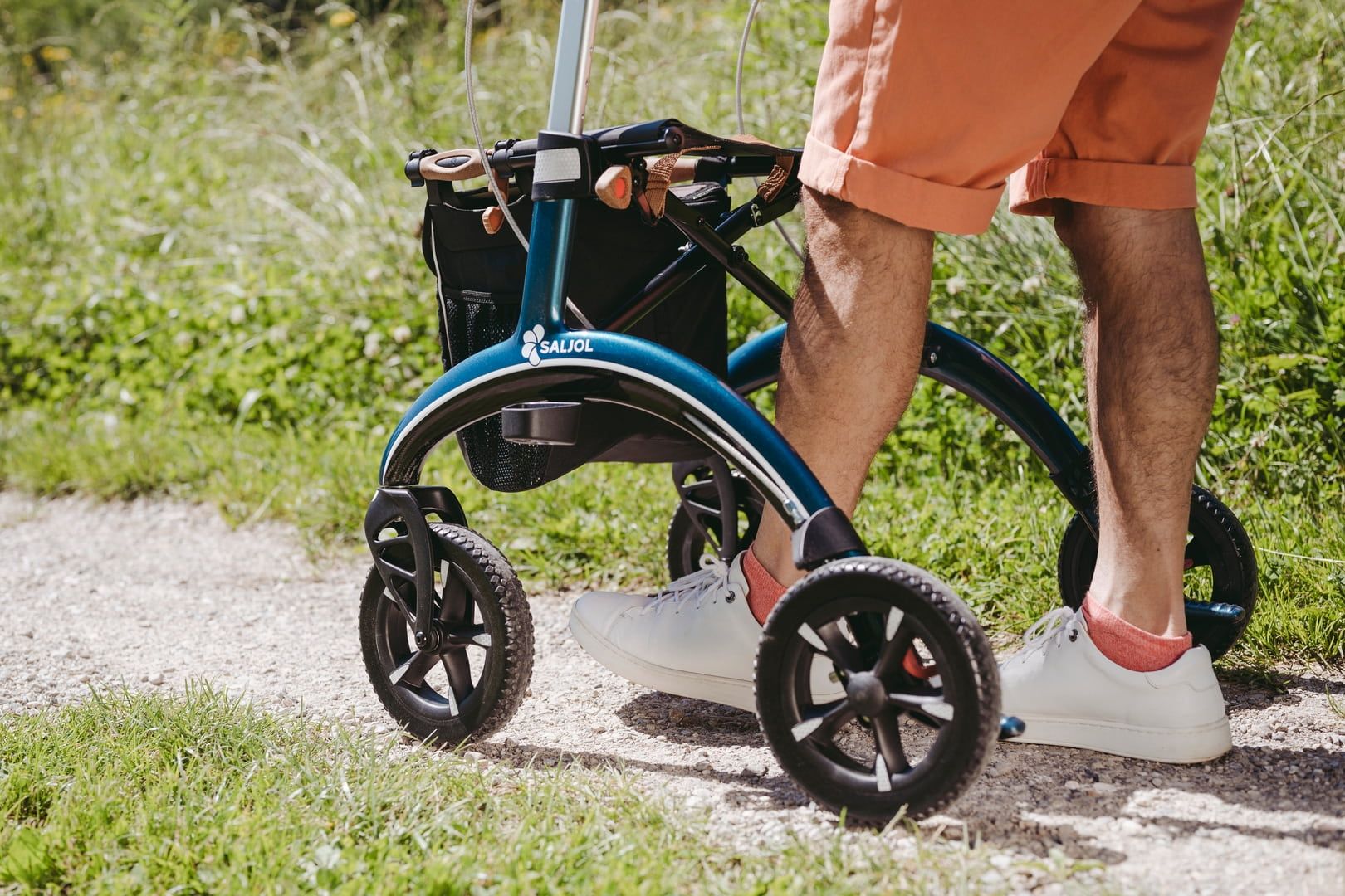 Person mit orangefarbener Hose und weißen Schuhen schiebt einen blauen Rollator auf einem Weg. Schwarze Räder.