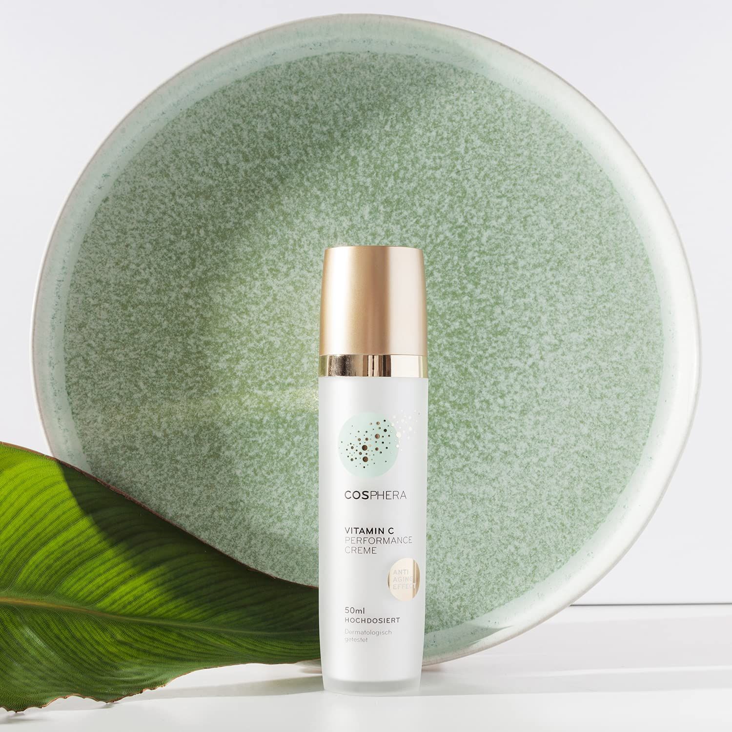 Flasche Cosphera Vitamin C Creme neben einem grünen Teller und Blatt. Pumpspender mit goldenem Verschluss.