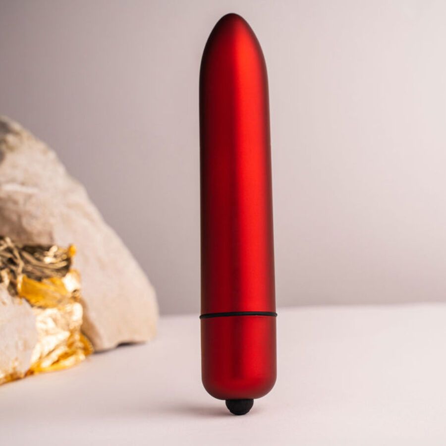 Roter Vibrator, zylindrisch, mit abgerundeter Spitze und schwarzem Knopf. Auf weißem Untergrund, neben Stein und Gold.
