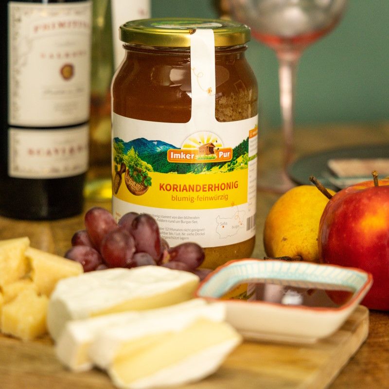 Glas mit ImkerPur Korianderhonig. Etikett mit Landschaftsmotiv und Schriftzug. Honig neben Käse, Trauben und Wein.