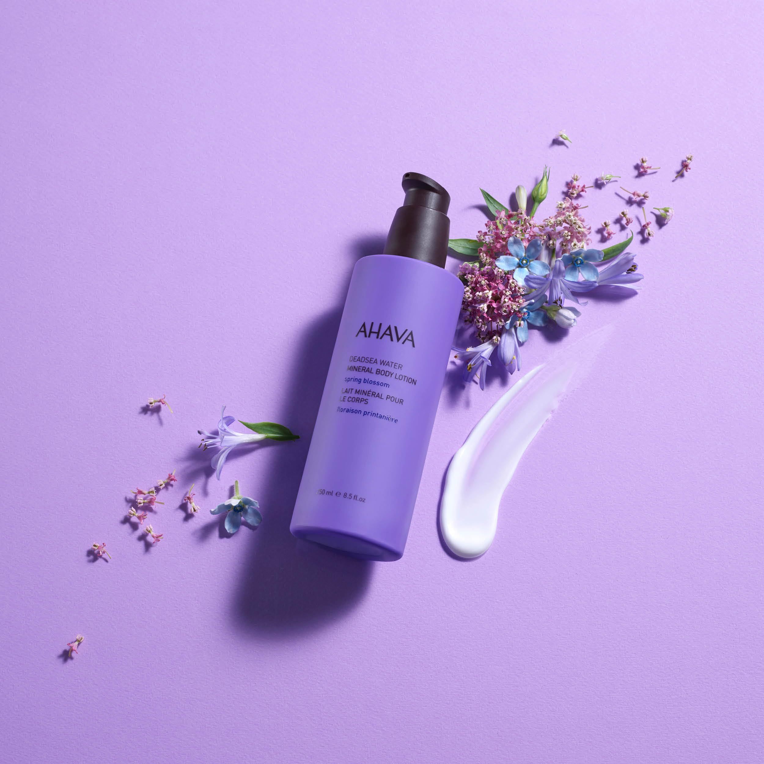 Lila AHAVA Mineral Body Lotion mit schwarzem Pumpkopf, umgeben von Blüten und einem Klecks Creme.