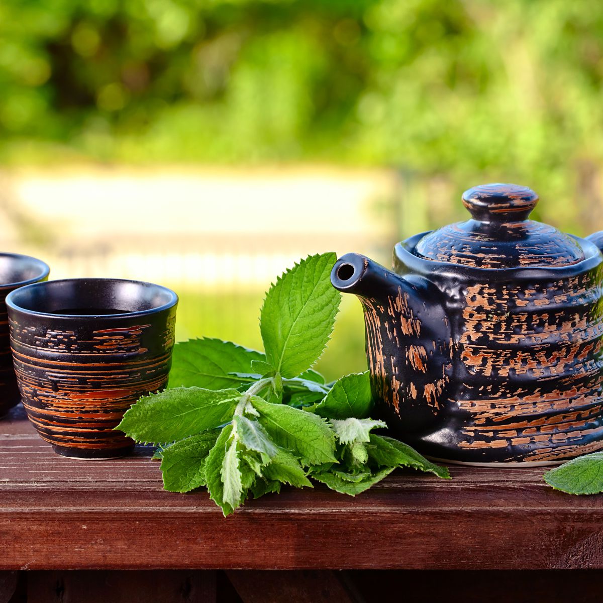 Teekanne und Tassen auf Holzoberfläche mit frischen Pfefferminzblättern. Hintergrund: unscharfer grüner Garten.