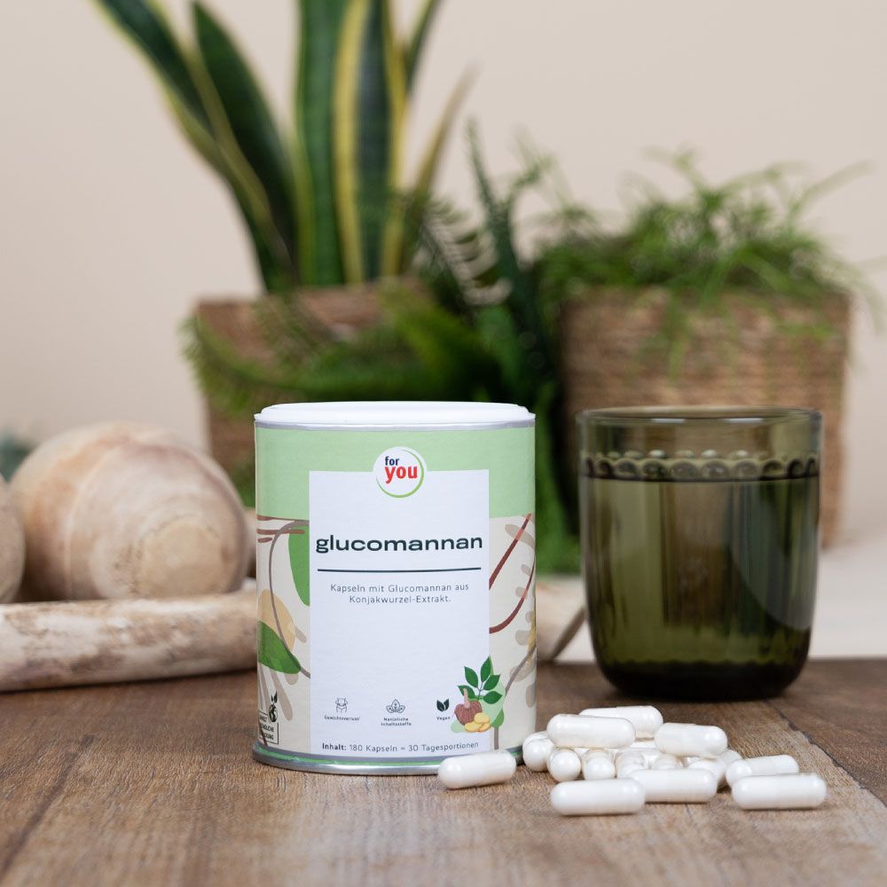 Dose "for you Glucomannan" auf Holzoberfläche. Kapseln daneben. Glas und Pflanzen im Hintergrund. Grün-weißes Design der Dose.