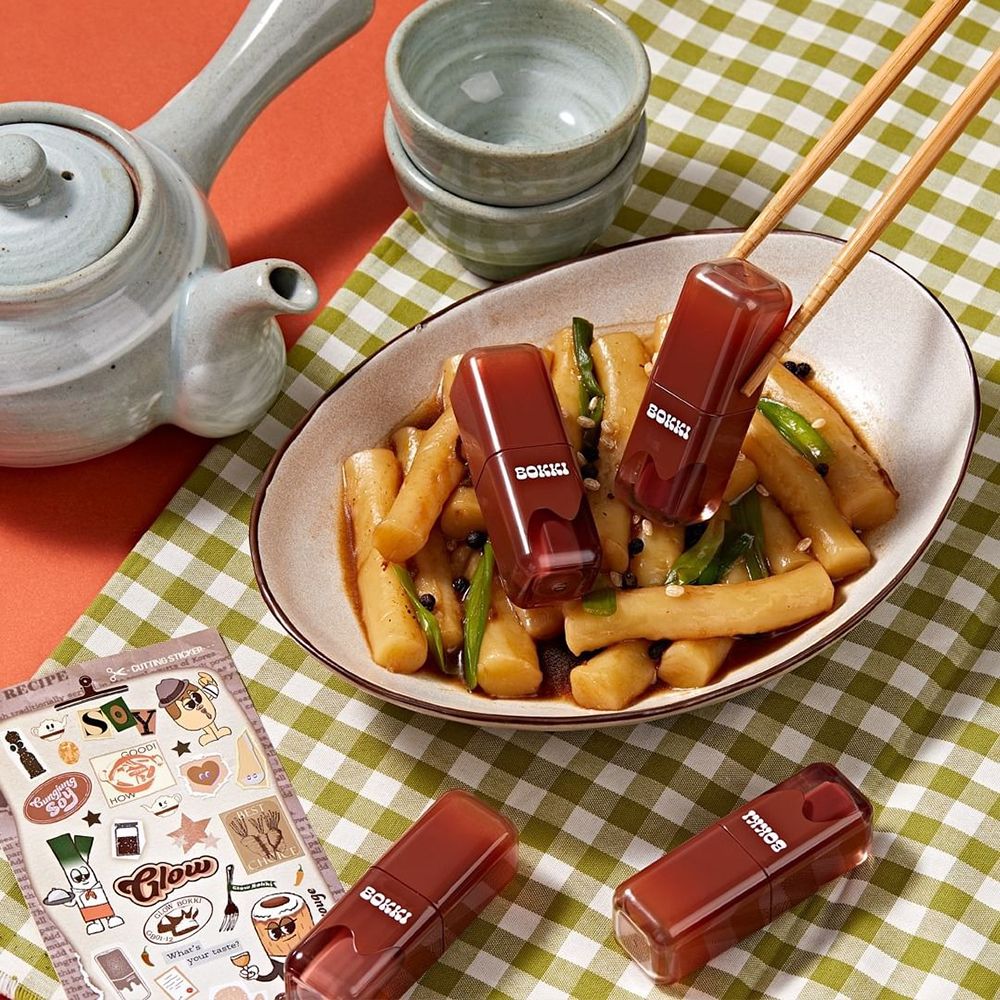 Drei Lippenfarben auf einem Tisch. Auf einem Teller mit Essen, daneben Stäbchen. Auf einem karierten Tischtuch.