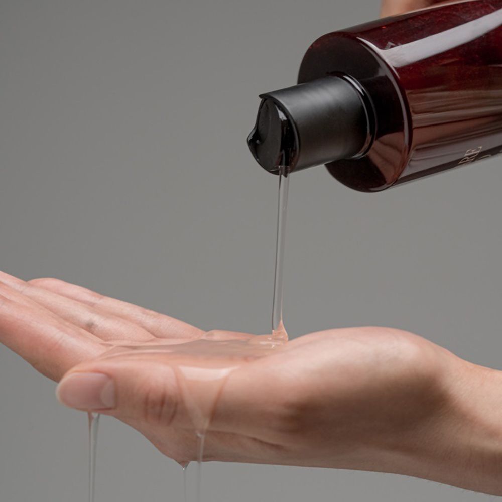 Hand gießt Shampoo aus einer braunen Flasche. Das Shampoo ist transparent.