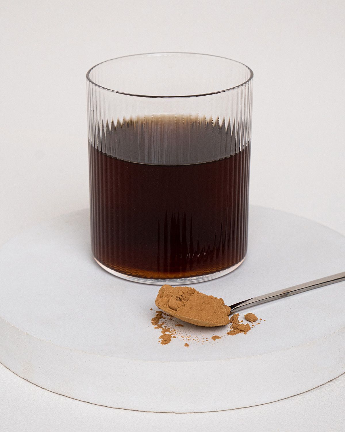 Glas mit Flüssigkeit und Löffel mit braunem Pulver auf weißer Oberfläche. Das Glas ist geriffelt.