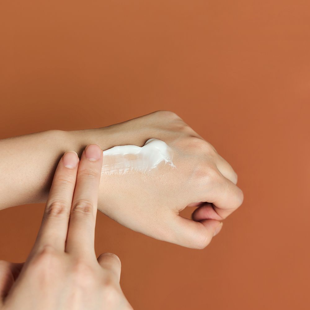 Hand mit Creme auf dem Arm. Creme wird mit zwei Fingern verrieben. Hintergrund: braun.