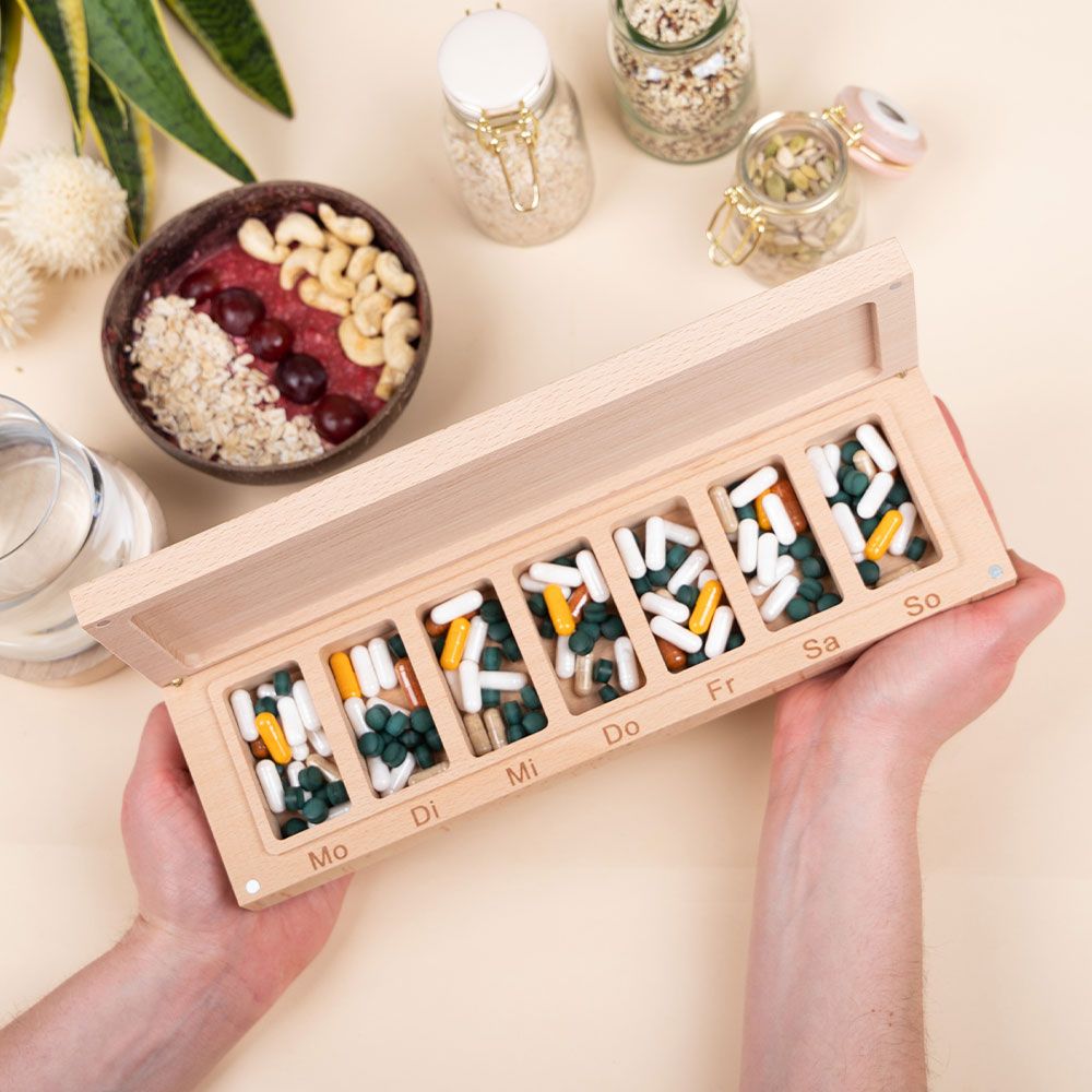 Tablettenbox aus Holz mit sieben Fächern, gefüllt mit Tabletten. Beschriftung mit Wochentagen. Geöffnet, in Händen gehalten.