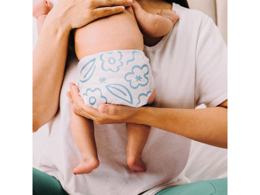 Baby mit Windel mit blauem Blumenmuster, gehalten von einer Person. Die Windel ist mit dem Produktnamen versehen.