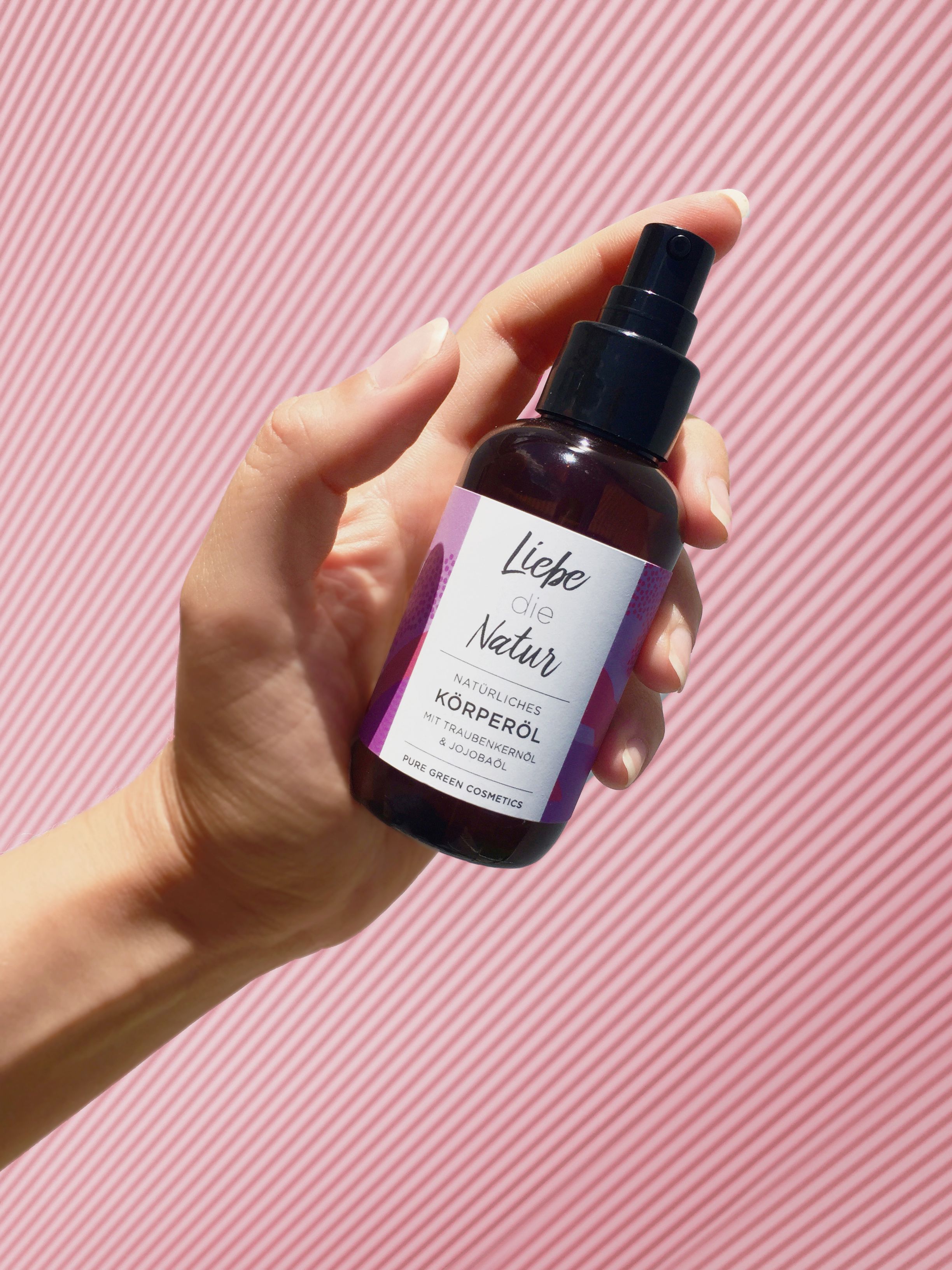 Braune Glasflasche mit Sprühkopf. Etikett mit Produktbezeichnung und Logo. Hand hält die Flasche vor rosa Hintergrund.