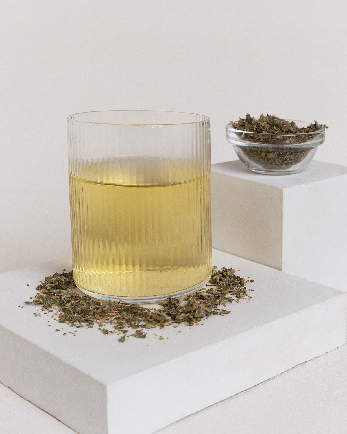 Glas mit Flüssigkeit und Kräutern. Kräuter auf weißem Podest und daneben in Schale. Flüssigkeit gelblich.
