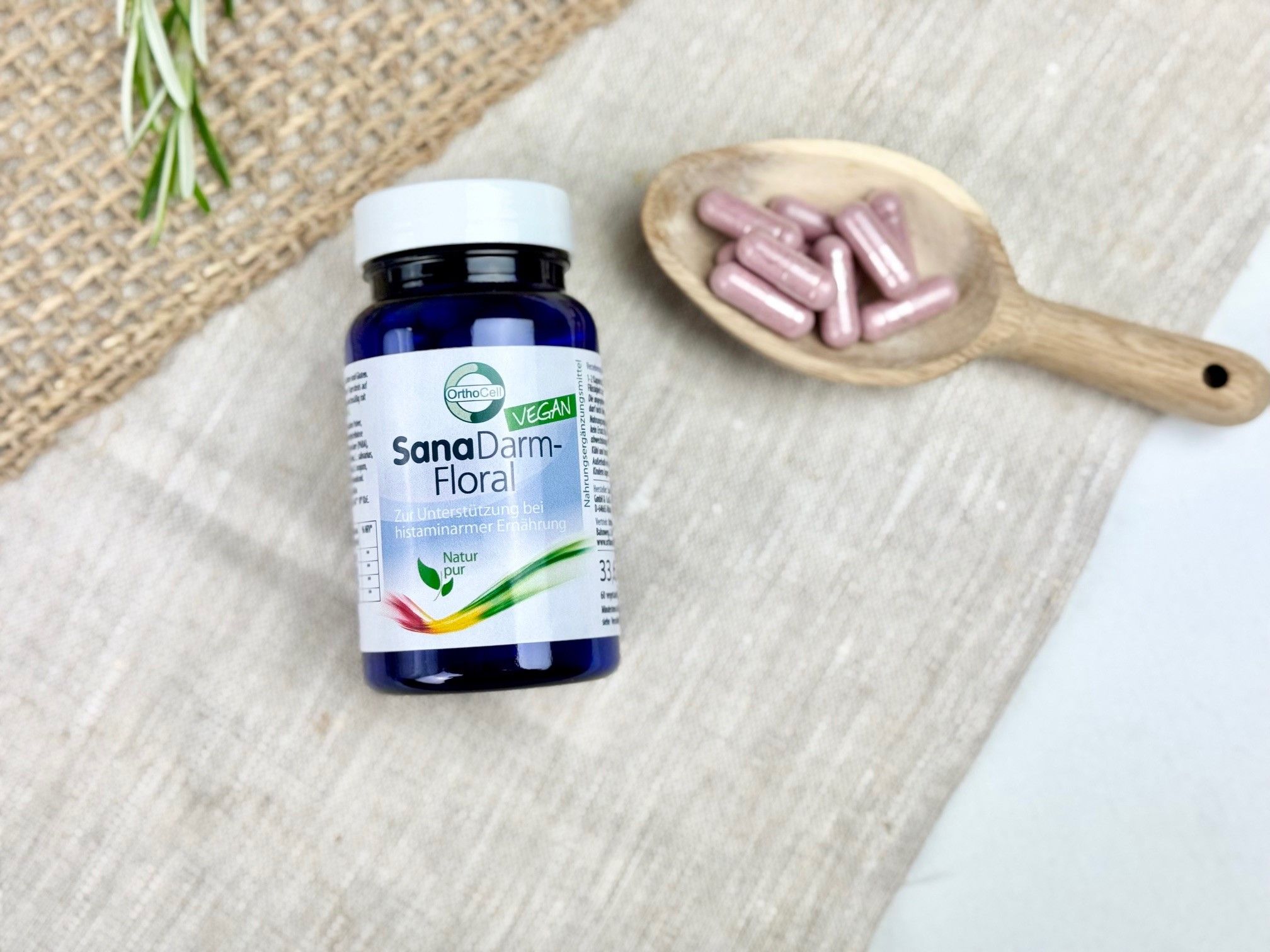 Blaue Flasche SanaDarm-Floral neben Holzlöffel mit Kapseln. Auf Leinenstoff. Vegan-Siegel. Rosmarinzweig.