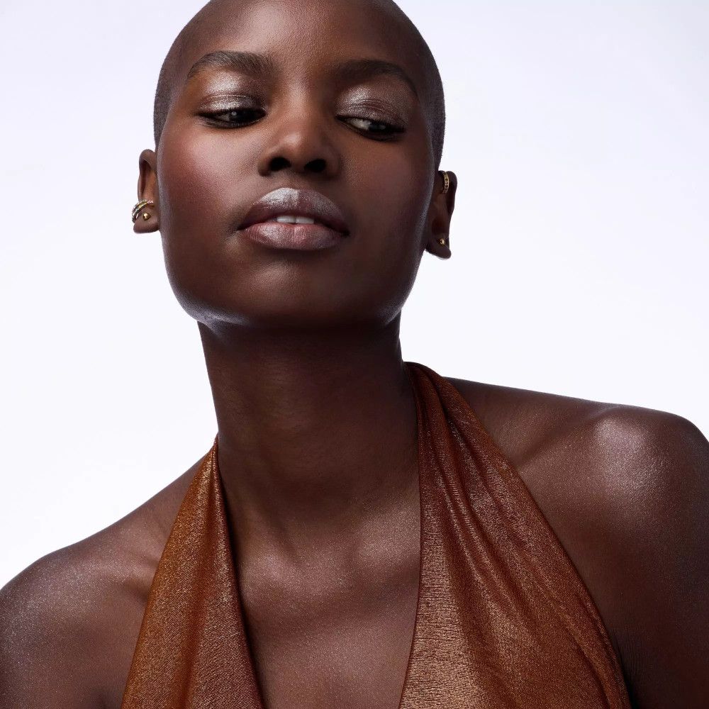 Porträt einer Frau mit rasiertem Kopf. Sie trägt ein braunes Oberteil und Ohrringe. Blick nach oben.