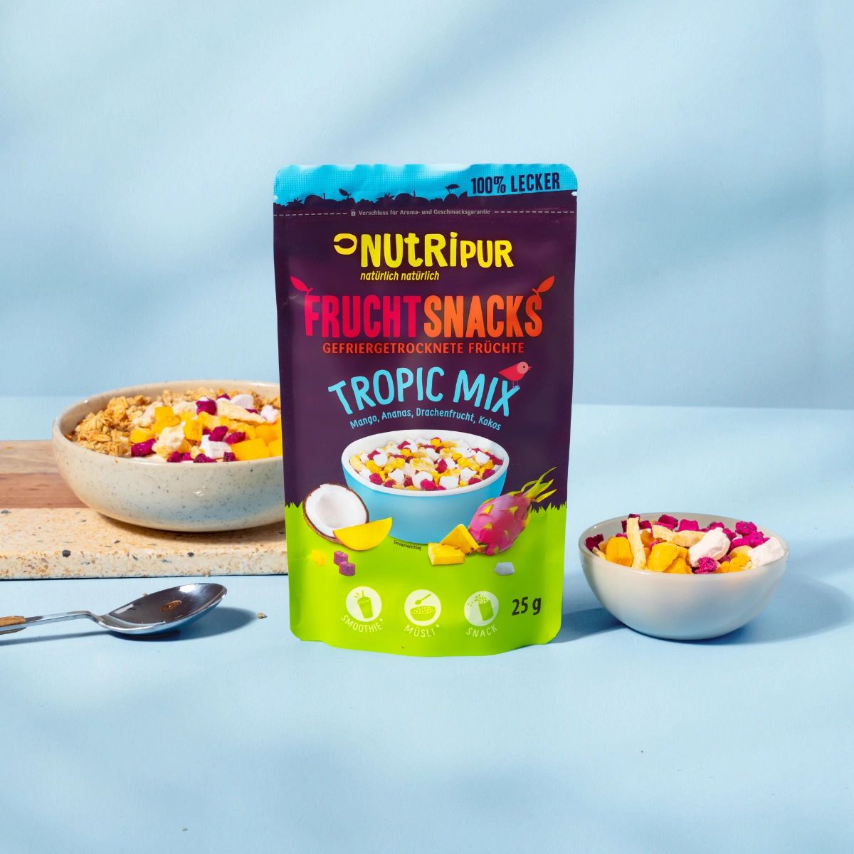 Beutel und Schale mit gefriergetrockneten Früchten. Aufschrift: NutriPur, Fruchtsnacks Tropic Mix. 25g. Auf blauem Hintergrund.