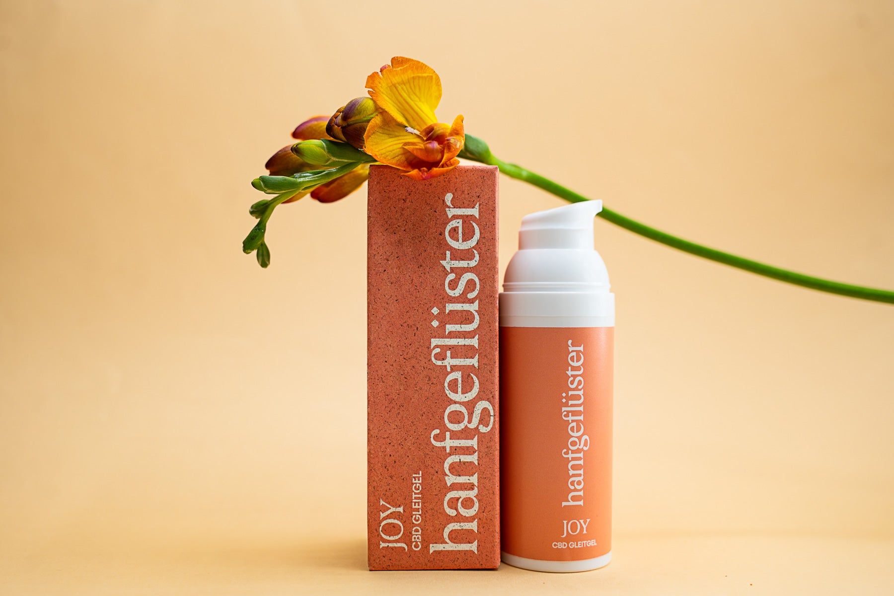 Orangefarbene Flasche und Verpackung mit weißer Schrift. Aufschrift: hanfgeflüster JOY CBD Gleitgel. Neben einer Blume.