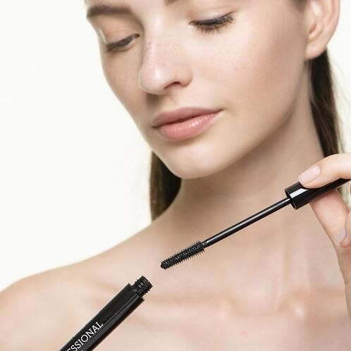 Frau hält Mascara-Tube und Bürste. Schwarze Tube mit Schriftzug. Bürste wird herausgezogen.