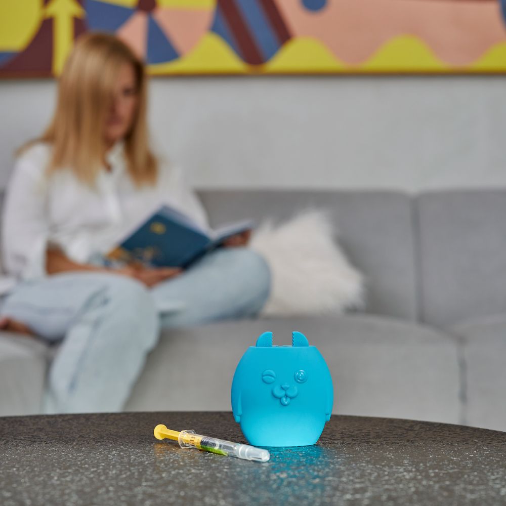 Blaues Medizinprodukt mit Spritze auf Tisch. Frau liest Buch im Hintergrund. Spritze mit gelber Flüssigkeit.