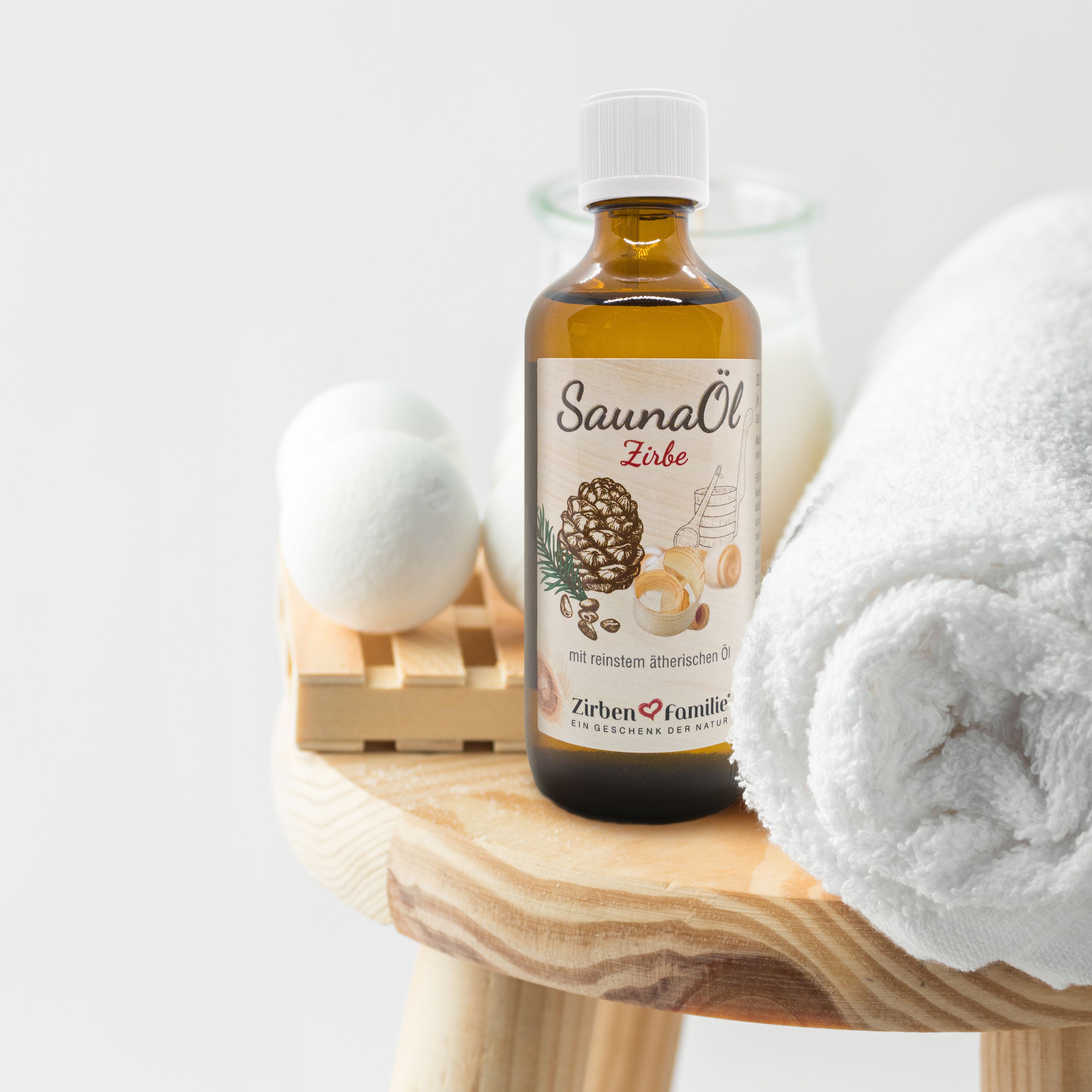 Braune Glasflasche mit weißem Deckel. Etikett SaunaÖl Zirbe. Auf Holzschemel mit Handtuch, Badesalz und Schwamm.