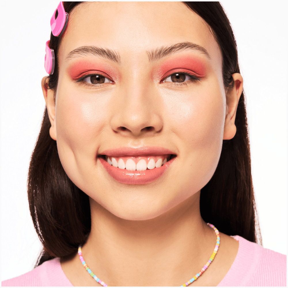 Porträt einer lächelnden Frau mit rosa Lidschatten. Sie trägt rosa Haarspangen und eine Halskette mit bunten Perlen.